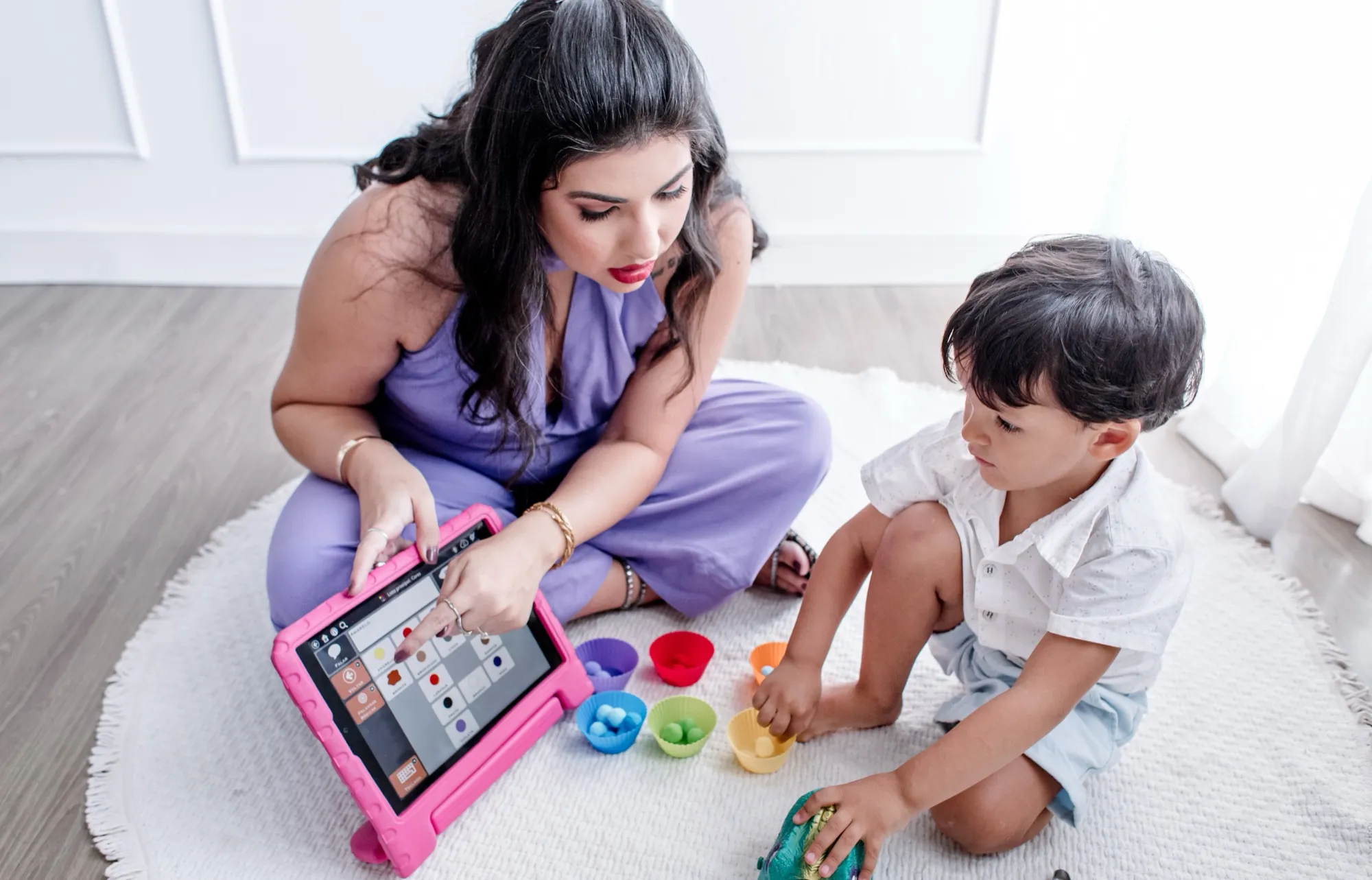 Izabella Souza aponta para um tablet com pranchas de comunicação alternativa enquanto um menino a acompanha, sentados no tapete