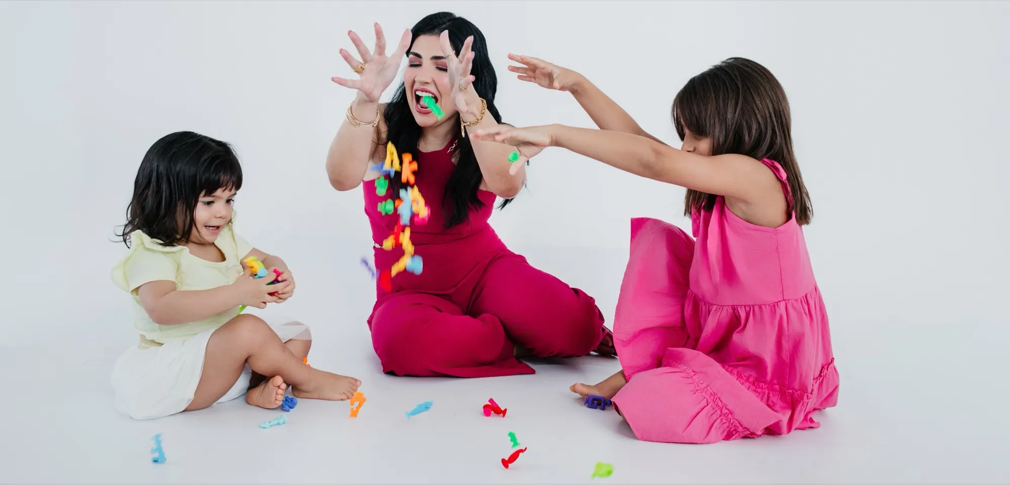 Izabella Souza sentada no chão brincando com duas crianças, entre letras coloridas espalhadas pelo chão